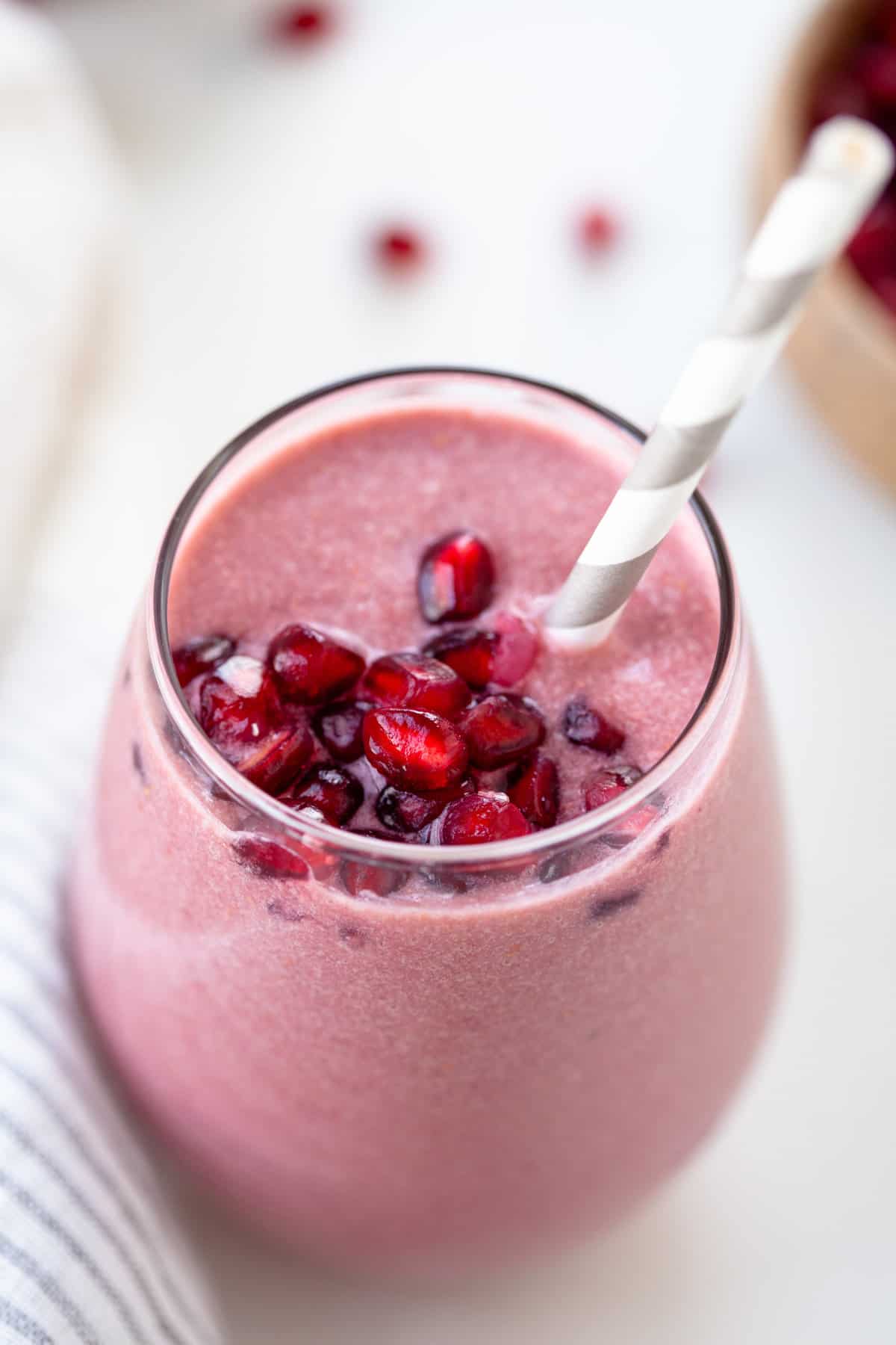 Pink smoothie in a glass with pomegranate seeds on top and a grey and white straw.