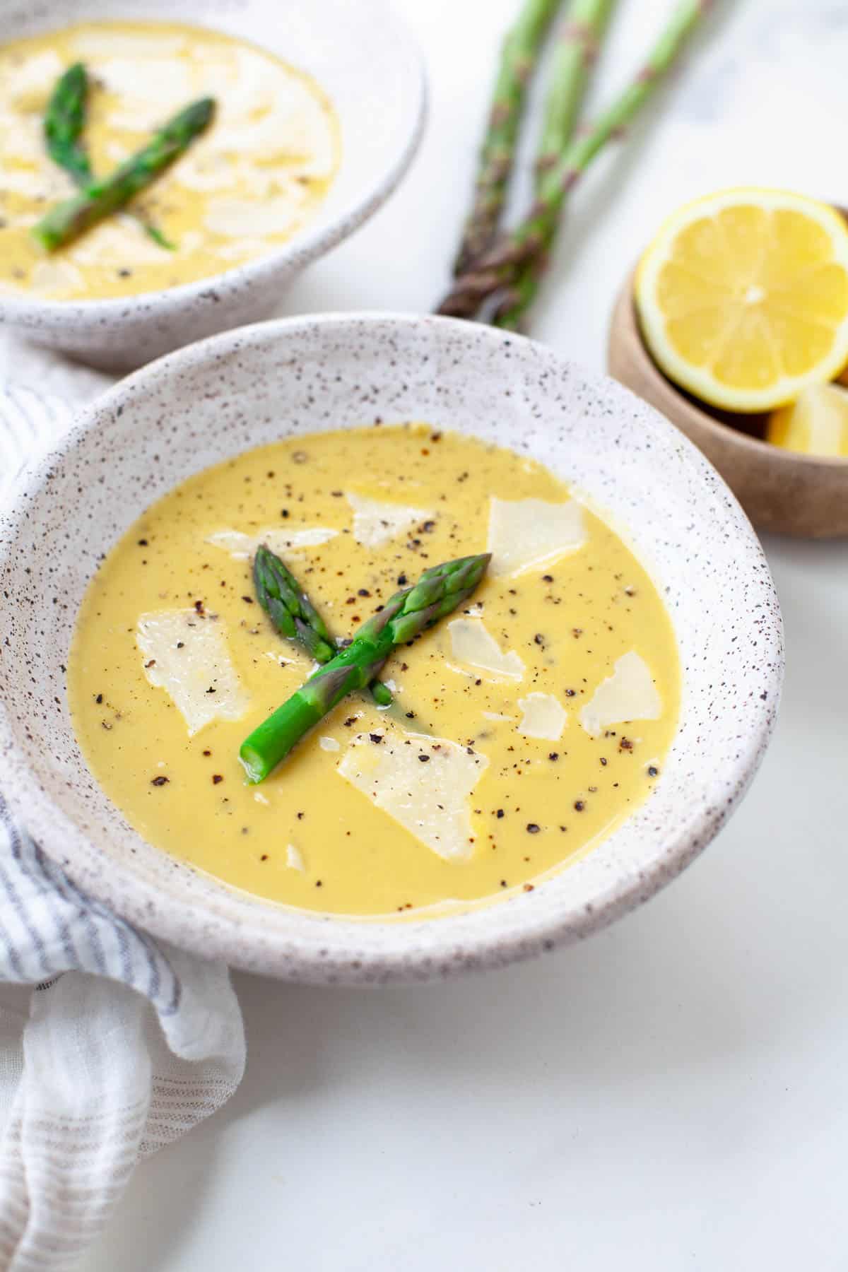 cream of asparagus soup in a white speckled bowl with asparagus pieces and lemons