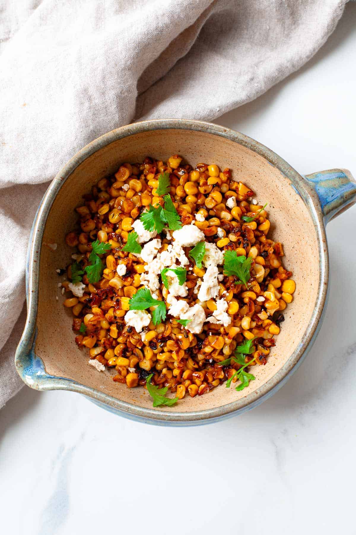 blackened corn in bowl with handle topped with fresh cilantro and crumbled feta cheese beside kitchen towel