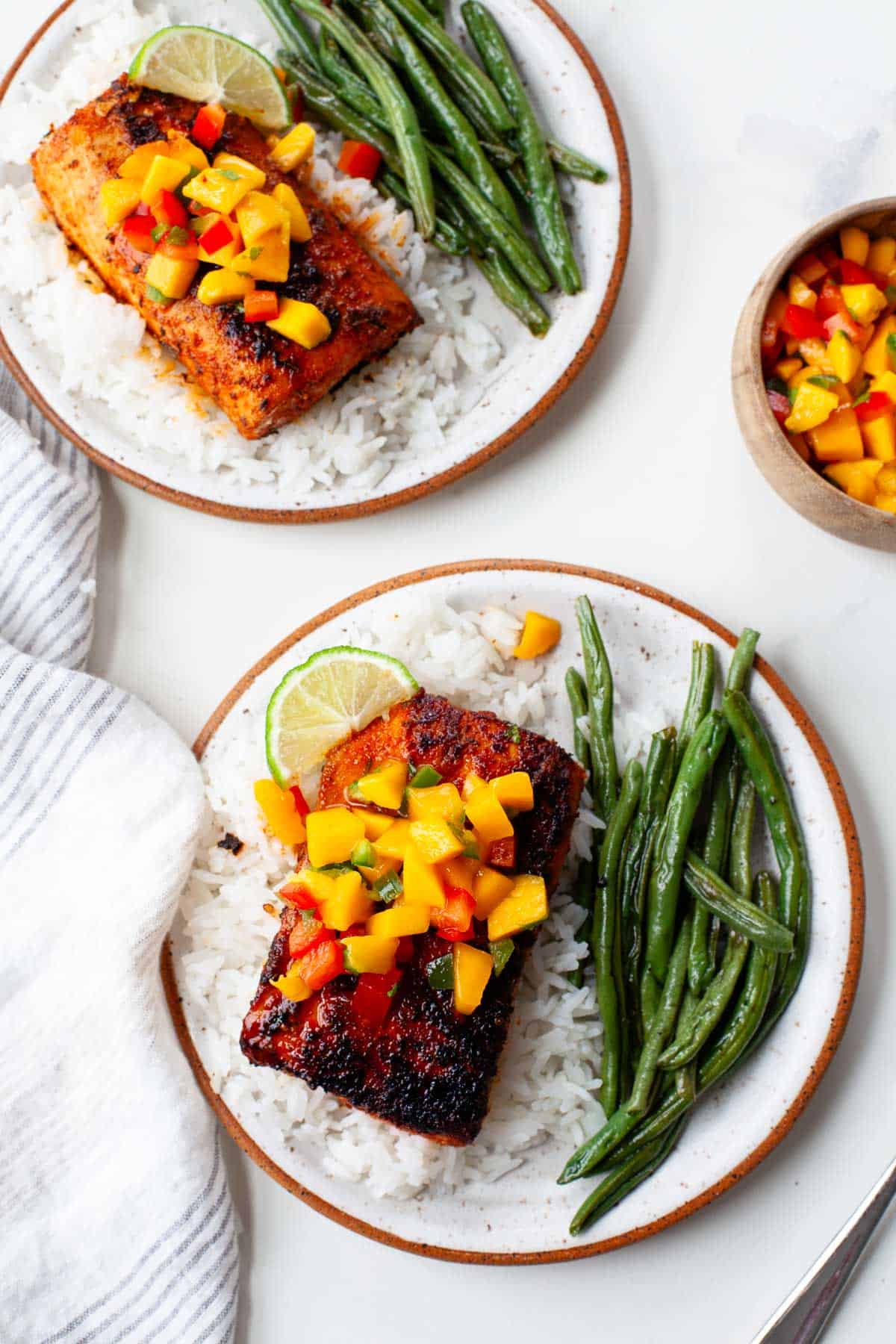 two plates of blackened mahi mahi atop beds of white rice, garnished with fresh mango salsa and served with green beans