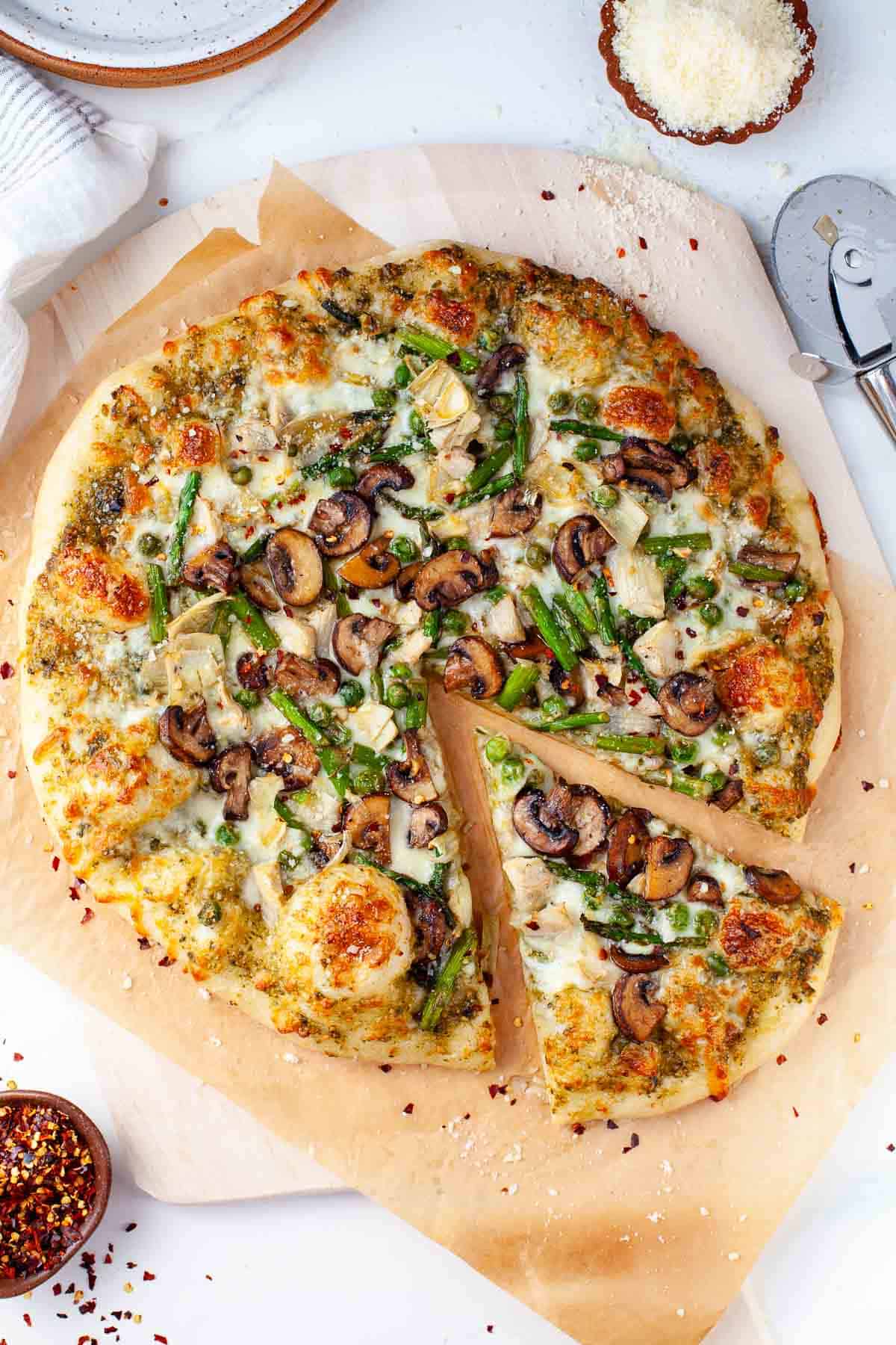 spring pizza with one slice cut on top of pizza stone that is beside a small bowl of grated parmesan cheese and a pizza cutter.